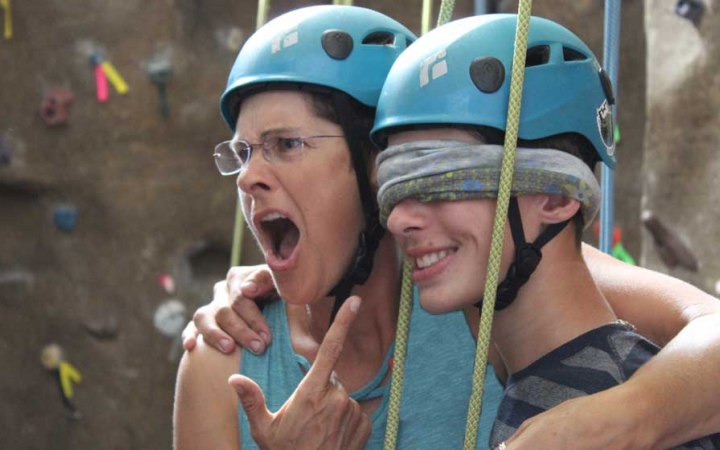 Two people who appear to have been rock climbing, based on their helmets and the rock wall in the background, embrace each other. One of them is blindfolded, and the other points, seeming impressed. 